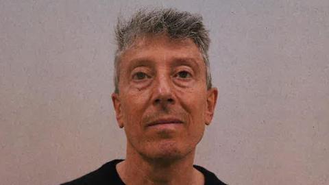 A custody mugshot of Howard Phillips, who has grey hair and no expression on his face. He is wearing a black T-shirt in front of a fuzzy grey background.