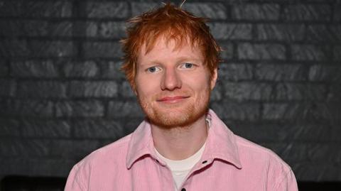 Ed Sheeran, looking straight at the camera, with short messy ginger hair, a ginger beard. He is smiling and is wearing a white T-shirt and pink jacket. He is standing in front of a black brick wall. 