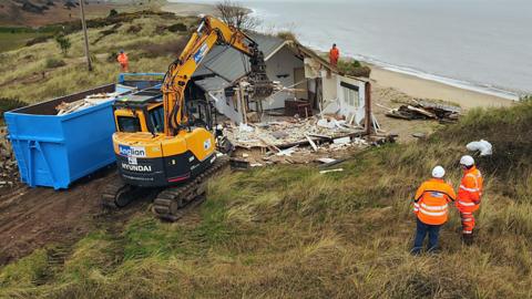 A house is partially demolished next to the cliff edge with a bright yellow digger next to a blue skip.