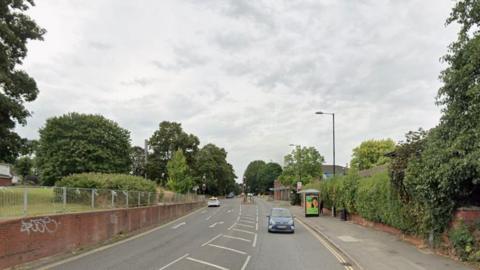 Cars travelling in both directions on a main road. Road markings, signs and traffic lights can be seen in the background. A brick wall and silver railings line one side of the road.