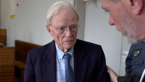 Iain Wares is wearing a navy suit jacket, blue checked shirt and blue tie. He has white hair and glasses. A man is placing a hand on his shoulder as they speak. A court of police officer can be seen in the background.