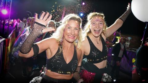 Two women in sparkly bras wave at the camera. It is night time.