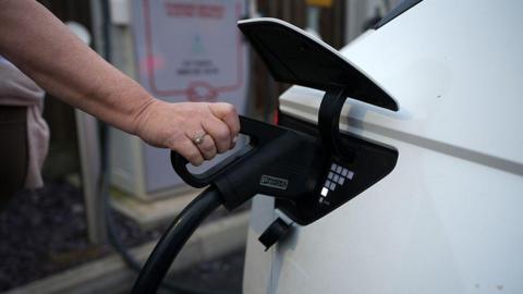 A close up of a person's hand holding a charger for an electric vehicle and putting it into the socket of a white car. In soft focus in the background, a charging point is visible.