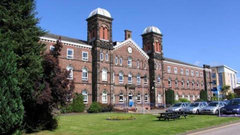 The Fusehill Street Campus in Carlisle. The large red-brick building has two towers either side of the main facade, with a wing extending to the side of both towers. There is a parking area, some grass and a bench in front of the building.