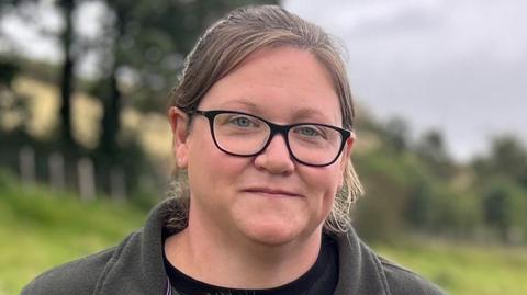 A woman with black rimmed glasses and a brown fleece standing in a field