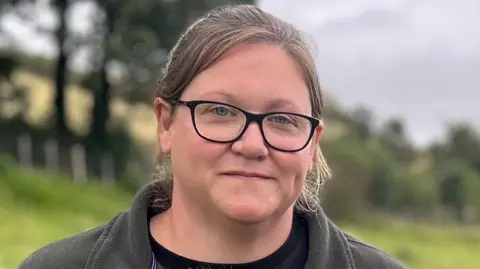 A woman with black rimmed glasses and a brown fleece standing in a field