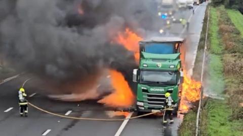 A green lorry is on fire as two firefighters are putting it out. There are big flames and a black plume of smoke.