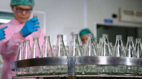 Employee inspecting glass bottles. They are wearing pink protective hear and blue glove.