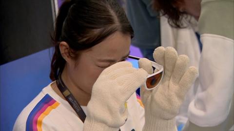 A woman with dark hair and white top with blue, purple and orange stripes on the sleeves tries out gloves and glasses intended to show what life is like for a dementia patient