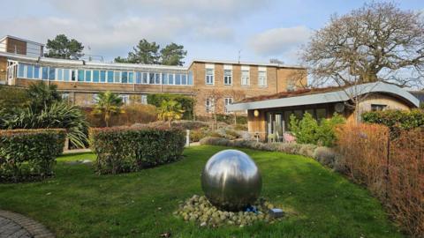 A modern building in the distance overlooks a green lawn and shrubbery. There is a wooden building to the left and a large stainless steel sphere in the middle of the grass.