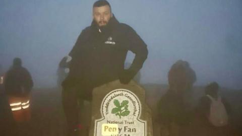 Lee, wearing a black coat, at the summit of Pen y Fan. He has his hand on a stone marker which marks the mountain's height at the top.