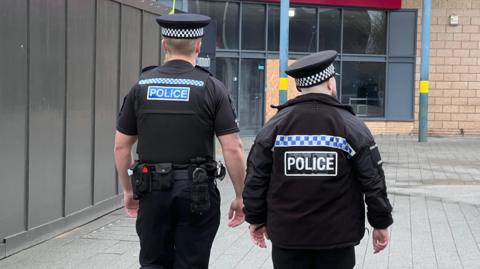 Two police officers on patrol in a pedestrianised area. They have their backs to the camera and are wearing their police caps and uniforms.
