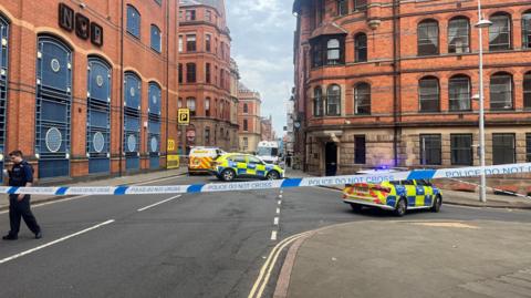 Police cordon tape and an officer in the foreground, with two police cars and a police van in the background, within the cordon.  