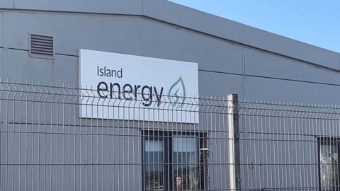 A grey, plastic-clad industrial building with an Island Energy sign and logo on it. The logo has a mint green leaf icon with a power lightning flash in the middle. In front of the building is a high metal fence.