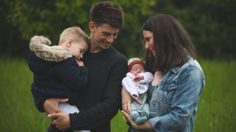 Sophie and her husband holding their two boys in their arms in a field smiling at each other