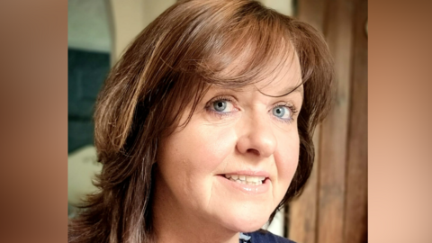 A picture of Lorraine Clements head, with her smiling at the camera. She has shoulder length brown hair, a blue jumper and is wearing 