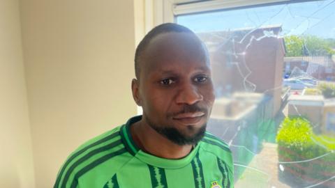  Mutasim Mohammed. He has short black hair and beard. He is standing in front of a smashed window on his home. He is wearing a Northern Ireland football top