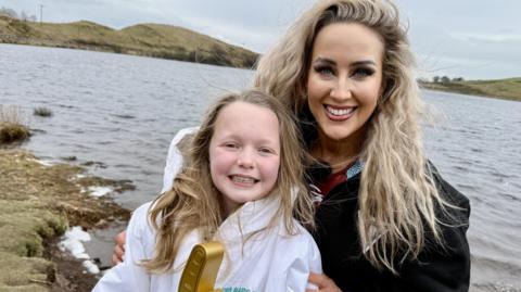 Gladiator Sabre and Grace stand by a loch. Grace can be seen holding her Glads of Honour award - a gold pointing finger statuette