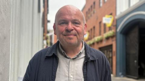 A bald man with grey stubble, he is smiling towards camera, wearing a grey shirt and navy jacket. The background is out of focus, red bricked buildings in Belfast's Cathedral Quarter
