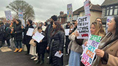 A group of men, women and children all wearing coats are stood at the side of the road holding placards