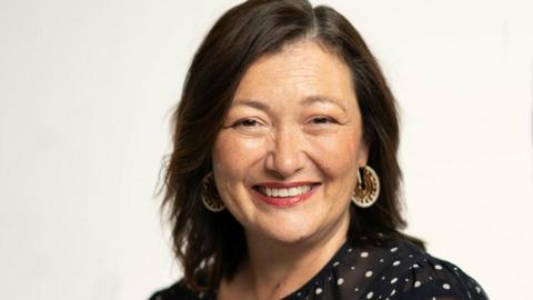 Caroline Miller pictured smiling with shoulder length brown hair, brown eyes and red lipstick. She is wearing gold hoop earrings and a black sheer top with white spots on it.