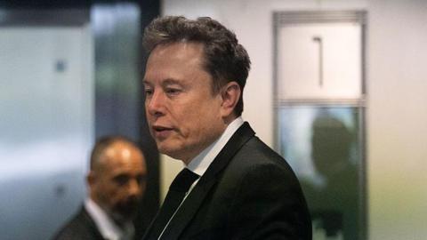 Profile shot of Elon Musk wearing a white shirt, black suit and black tie