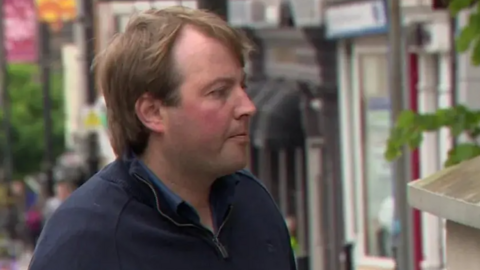 Jonathan Creswell with long brown hair wearing a navy zip-up fleece and polo shirt as he walks along a street.