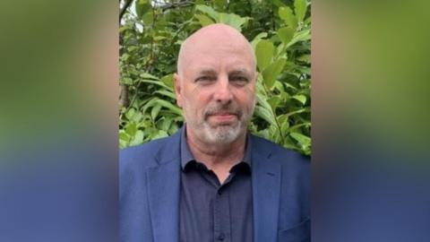 A man in a blue shirt and jacket standing outside in front of some greenery