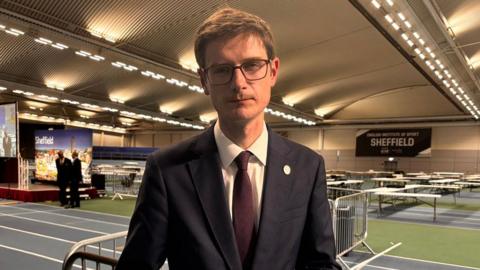 A man stands in a leisure centre where tables are in the background for the vote count. He has a navy suit, burgundy tie, brown short hair and black square glasses. He has a neutral expression but looks sad.