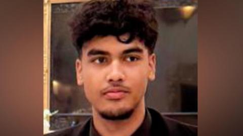 Absi Ahmed looking to the right of the camera. He has dark, curly hair which is short on the sides and long on the top. The photo is cropped close, with only his top and shoulders visible. He is wearing a black suit jacket, black shirt and black tie.