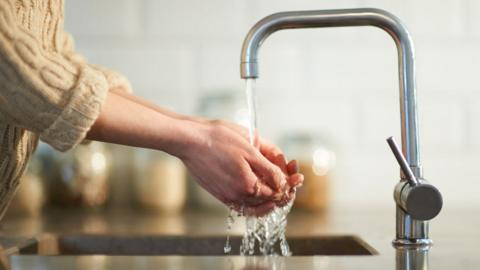 A woman holding her hands under water running from a silver tap in a sink in a kitchen