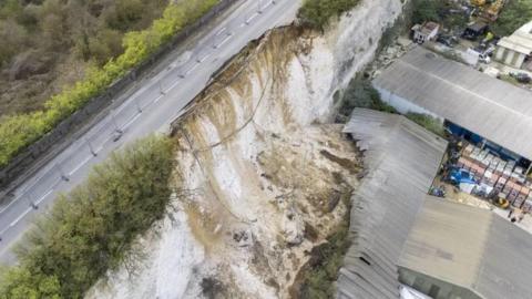 A road along a chalk clifftop with industrial buildings underneath. The road has partly fallen away after a landslip.