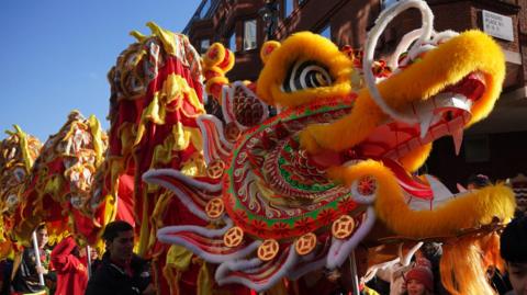 A close-up of a vibrant Chinese dragon costume with intricate patterns, fur being carried through the street by performers during a parade. Red lanterns hang in the background.