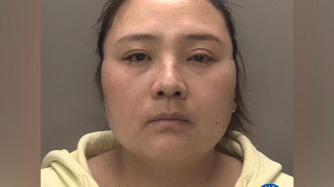 A mugshot of a woman who has dark hair and brown eyes, wearing a yellow hoodie