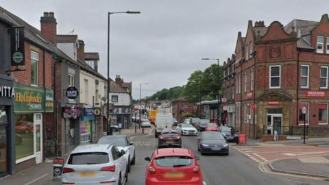 A road with shops on both sides and lots of cars travelling in both directions. Cars parked on both sides of the road and a bus stop on the left hand side.