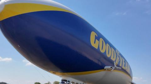 The blue-and-yellow Goodyear blimp moored on an airfield as people gather near the passenger cabin before or after a flight.