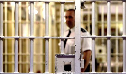 A prison guard stands behind a locked white metal gate inside HMP Pentonville. The gate is in focus with the guard and background blurred. The guard is dark trousers, a white short-sleeved shift and a black tie.