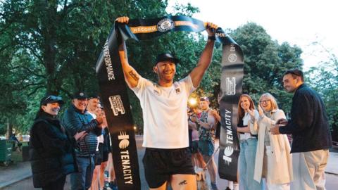 Fergus Crawley is wearing a light top, black shorts and dark cap with a closely trimmed beard and moustache. He is holding a banner which reads "Record Holder" and "Guinness World Records". There are trees behind him and people cheering around him.