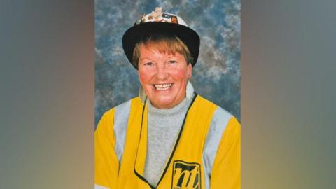 A portrait of Dot, smiling at the camera. She has short brown hair and is wearing a black and white hat with colourful stickers, and a yellow high-vis jacket. 