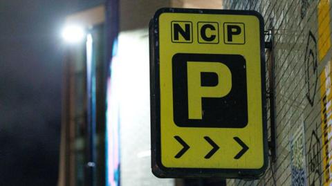 The logo of an NCP car park, which is rectangular, yellow and black, attached to a grey brick wall that has graffiti on it. It is dark and the background is blurred.