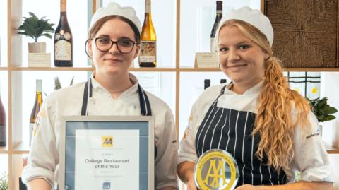 Edie (left) and Millie (right) wearing chef hats and holding the AA College Restaurant of the Year awards, with shelves of wine on display behind them.