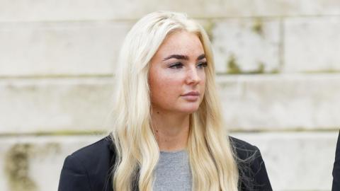 Close up of Megan looking away from the camera. She wears a grey t-shirt and a black blazer. Megan has long blonde hair and black eyelash extensions.