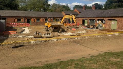 An old red brick stable block, with a digger in front of, where building work is underway. There is a large area of gravel in front with some grass closer to the front.