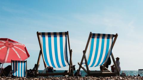 The back of two deckchairs and a red parasol on a beach under a blue sunny sky.