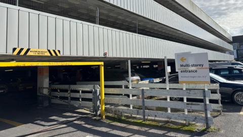 Entrance to the multi-storey section of Moorfield car park in Truro. A sign saying "multi-storey entrance" with a yellow arrow pointing left is outside the entrance, which is marked with a 1.9m maximum height barrier. Several cars are parked outside the multi-storey.