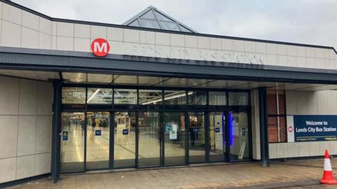 The entrance to a building which has a modern design with light-coloured panels and a triangular glass roof section. Above the glass doors, the sign reads ‘Leeds City Bus Station’.
