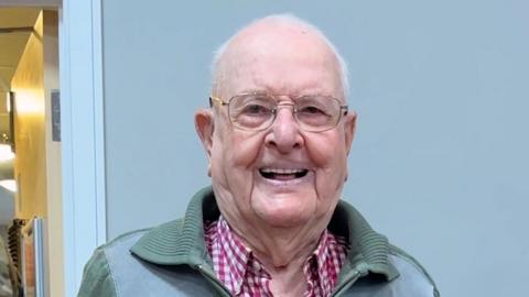Jack Waterfield smiling at the camera, with a glass of fizz in his hand. He has short white hair and glasses, and is wearing a green zip-up top over a white-and-red checked shirt. He is in a room with some people in military uniform around him.
