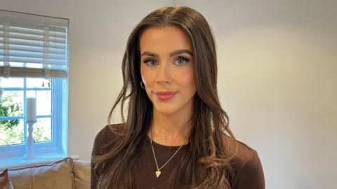 Charli Aitken - a woman - looks at the camera. She is standing in front of a sofa and a window. She has long brown hair and a brown top. She is wearing hoop earrings and a necklace with a pendant.