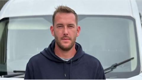 Toby Kennedy-Hall stares into the camera as he stands in front of his van. He wears a dark blue hoodie, with a grey T-shirt just visible underneath.