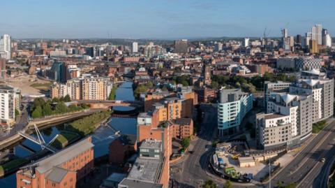 An aerial photograph of Leeds showing an urban skyline and a canal running through the middle. There is a mix of modern high-rise buildings and older brick structures.